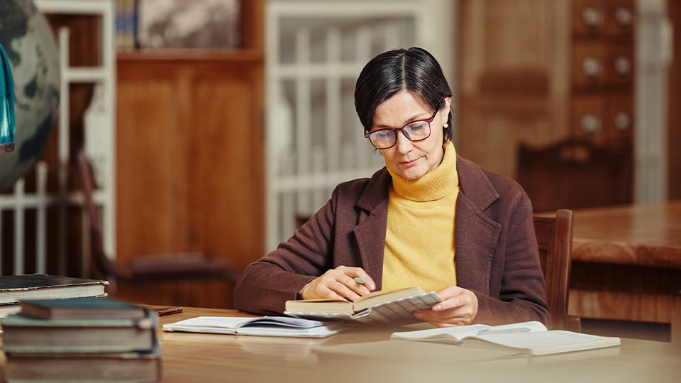 A woman reading a book at a table in a library setting, surrounded by books and wooden furniture. She is holding a pen.