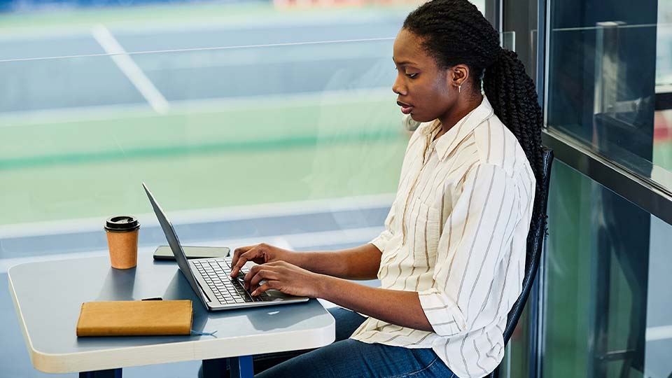 Business professional working on a laptop at a desk, with a notebook and coffee cup, overlooking a sports field