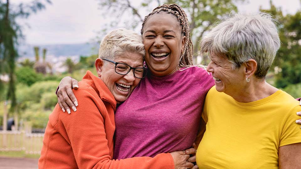 Three women in colorful clothes laugh and embrace in an outdoor setting