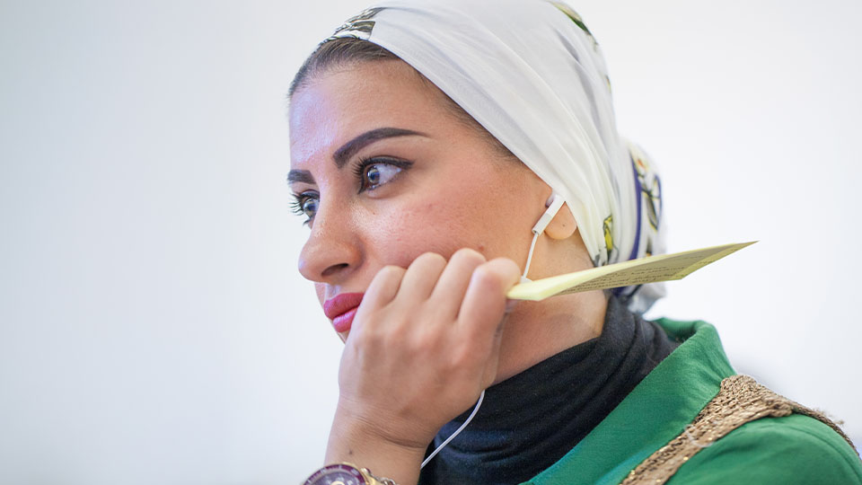 Woman in hijab with ear pod headphones holding a paper and looking at a screen