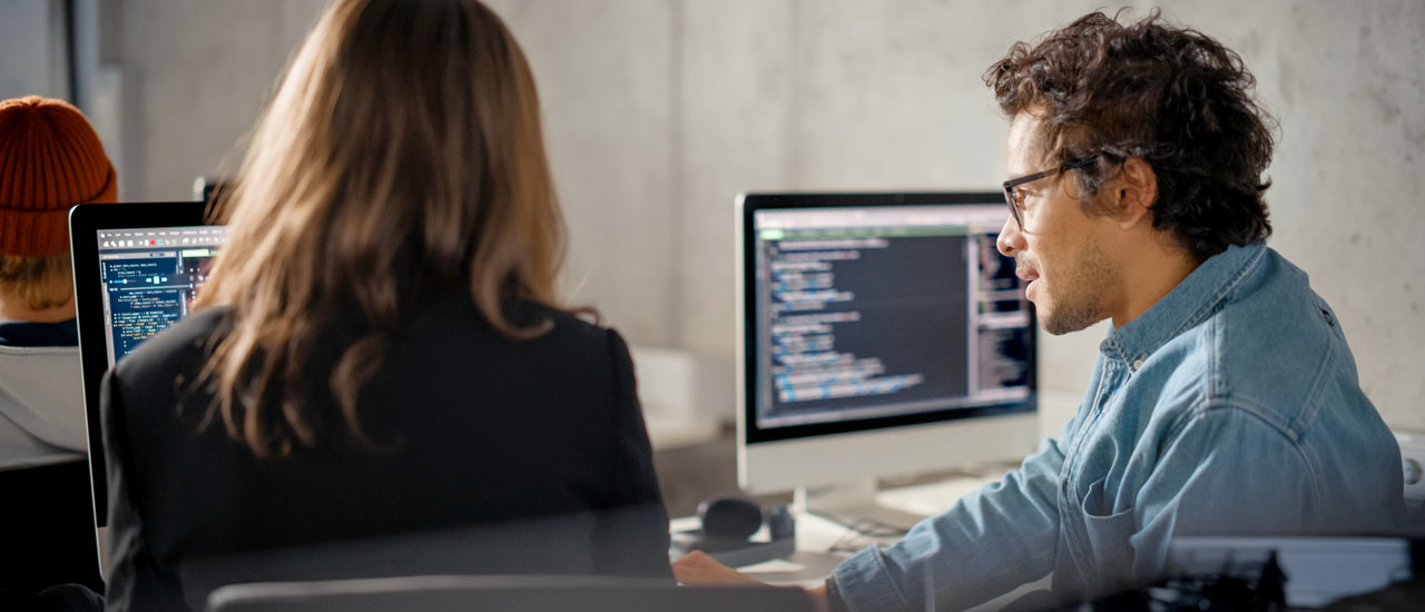 Two computer monitors display code, and a man and woman sit in front of one while discussing what's on screen