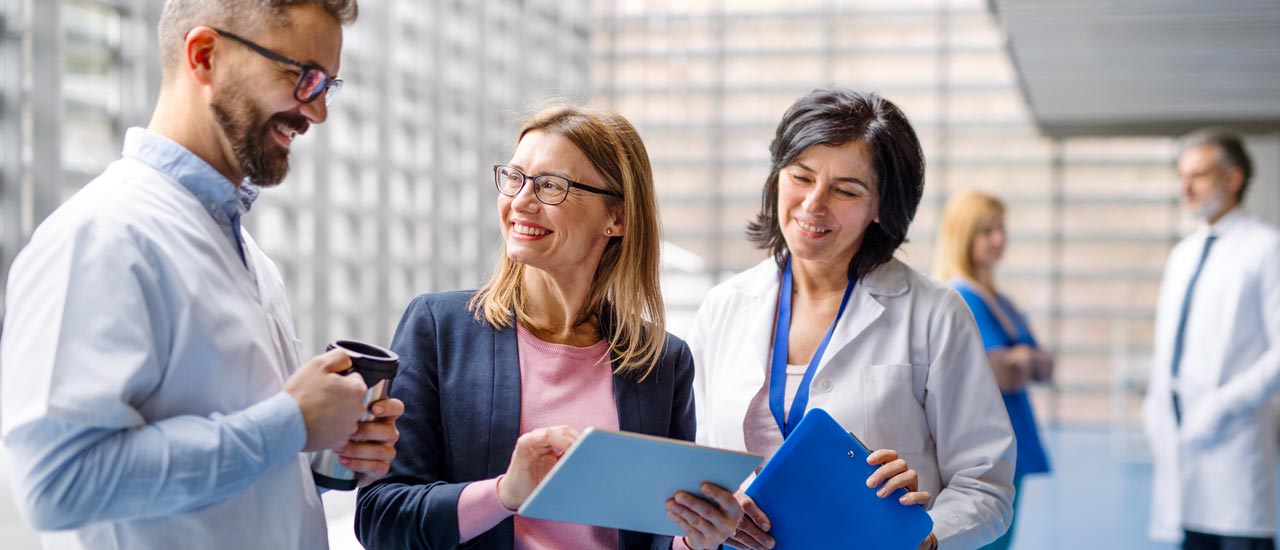 Woman in business attire holds a tablet and smiles. Two people in lab coats stand smiling on either side of her