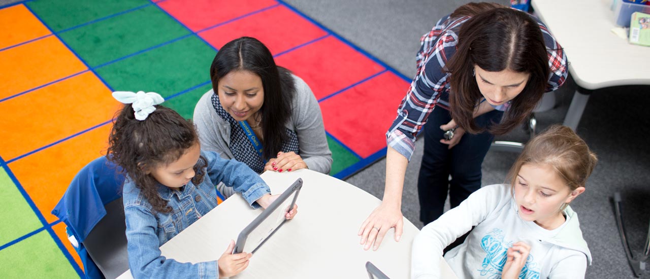 Teacher assisting young students seated around a classroom table as they use tablet devices, with a colorful rug in the background.