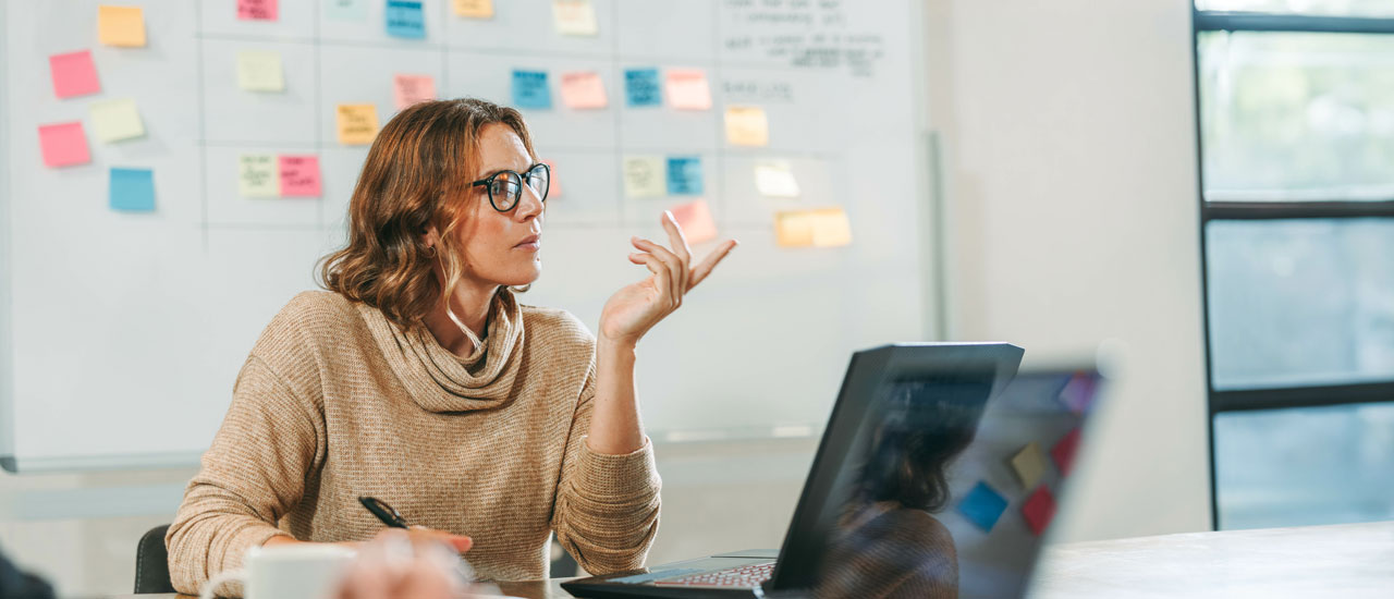 A woman looks engaged in a conference room with colorful sticky notes on a whiteboard in the background. Her laptop is open and she has a pen in her hand.